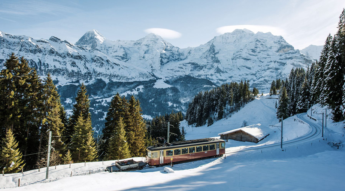 An old yellow and mountain train sits on snowy tracks with snow-covered mountains in the background